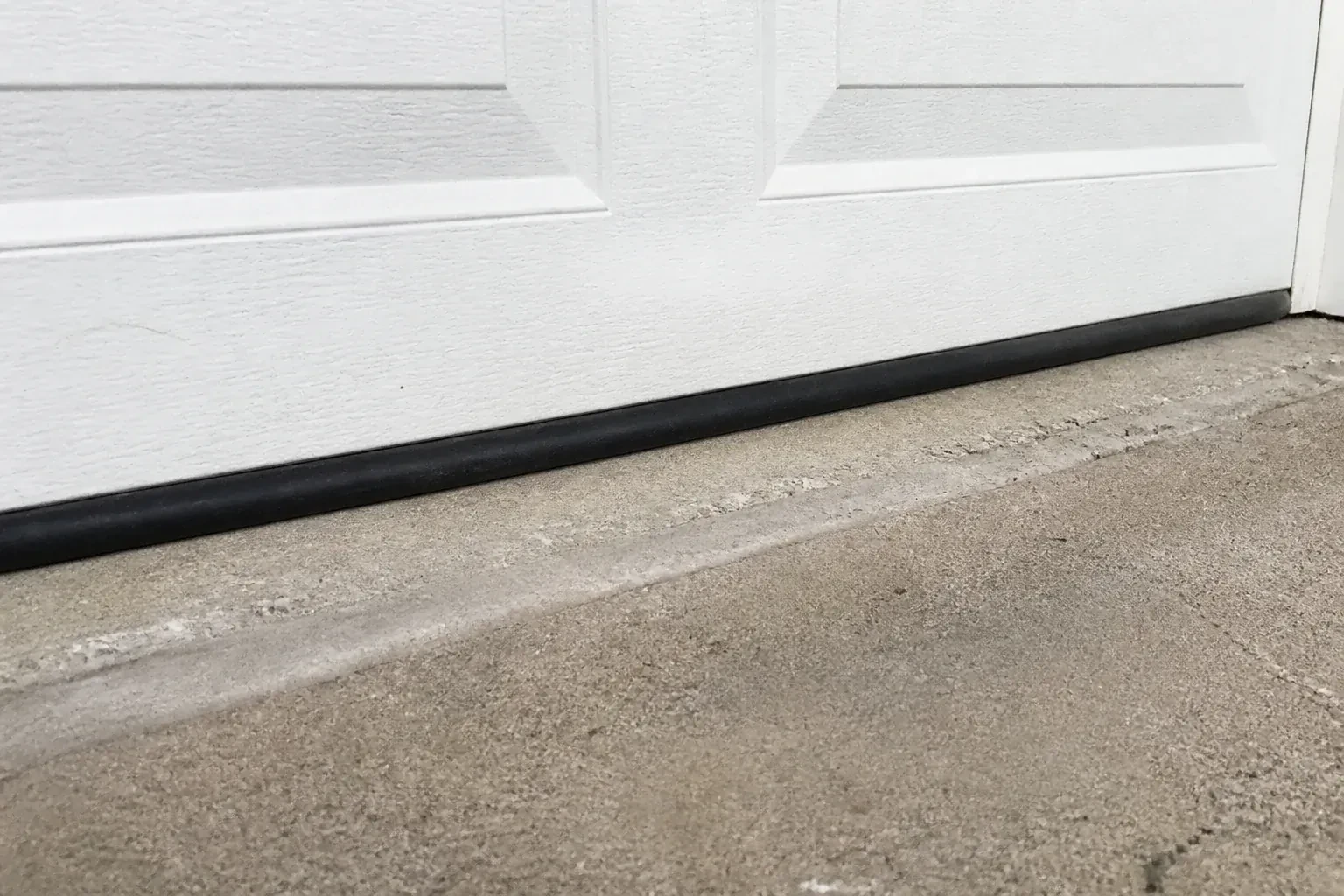 Black bottom seal along a closed white garage door at a concrete threshold. — Garage Door Bottom Seal Replacement, Whitchurch-Stouffville