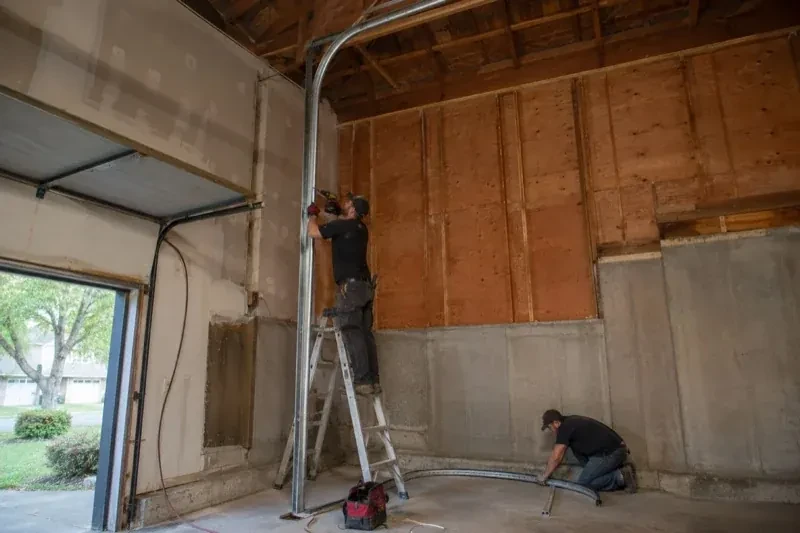 One person on a stepladder working beside a tall curved garage-door track while another kneels beside loose track sections in an unfinished garage. — High-Lift Garage Door Conversion, Whitchurch-Stouffville
